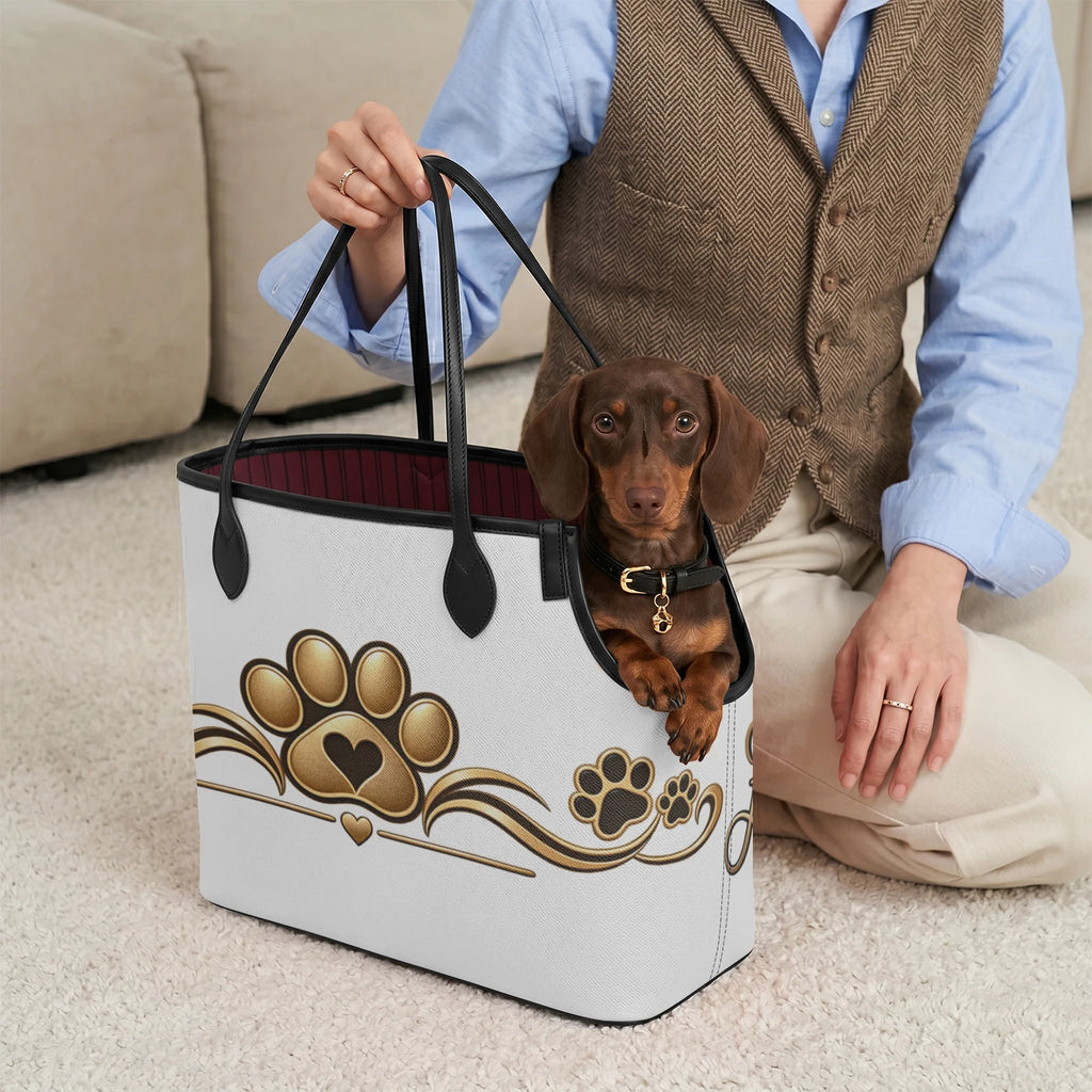 Brown dog peeking from the paw-print pet carrier tote beside a person seated indoors.